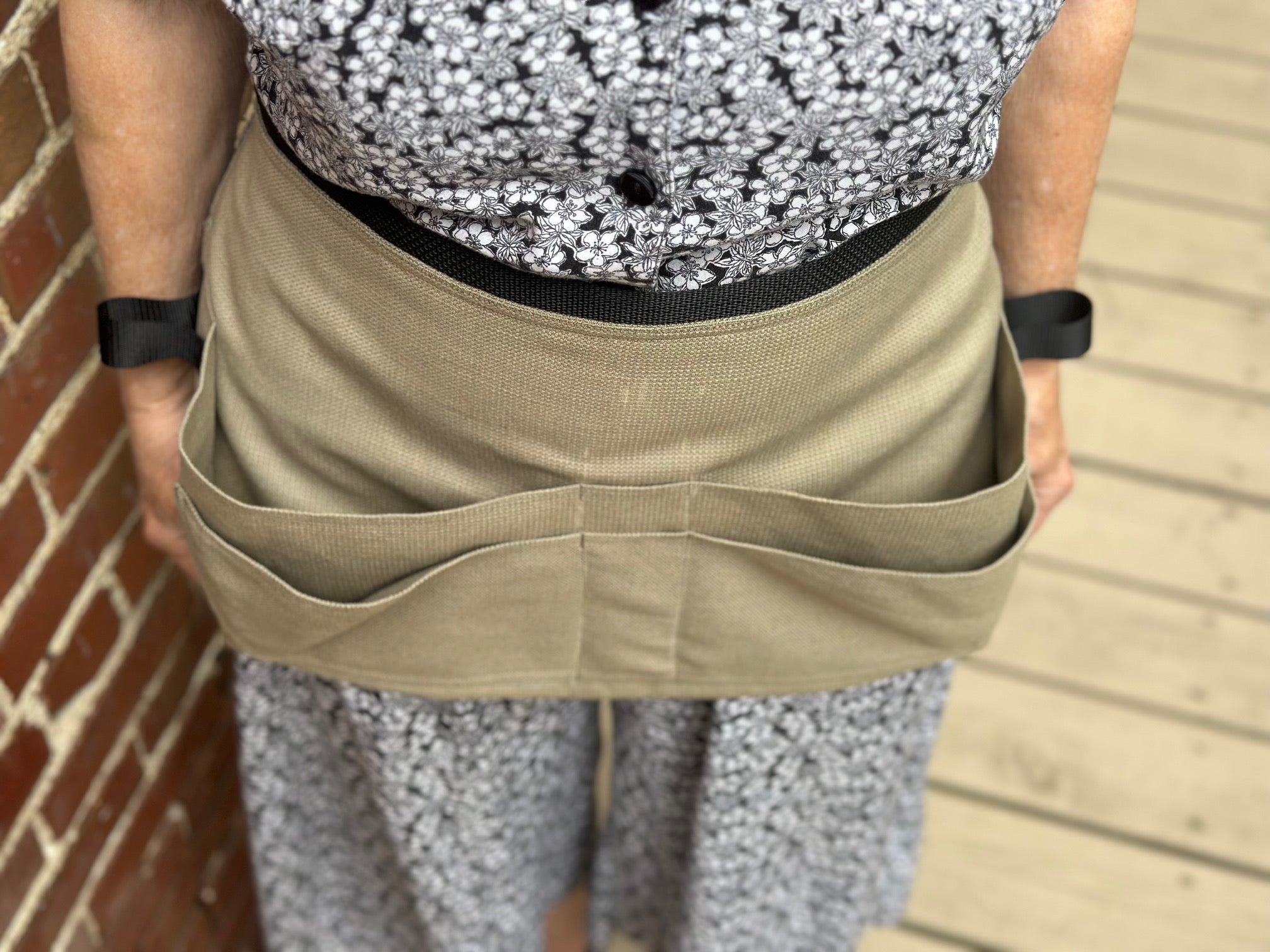 Person wearing a beige apron tool belt with multiple pockets against a brick wall.