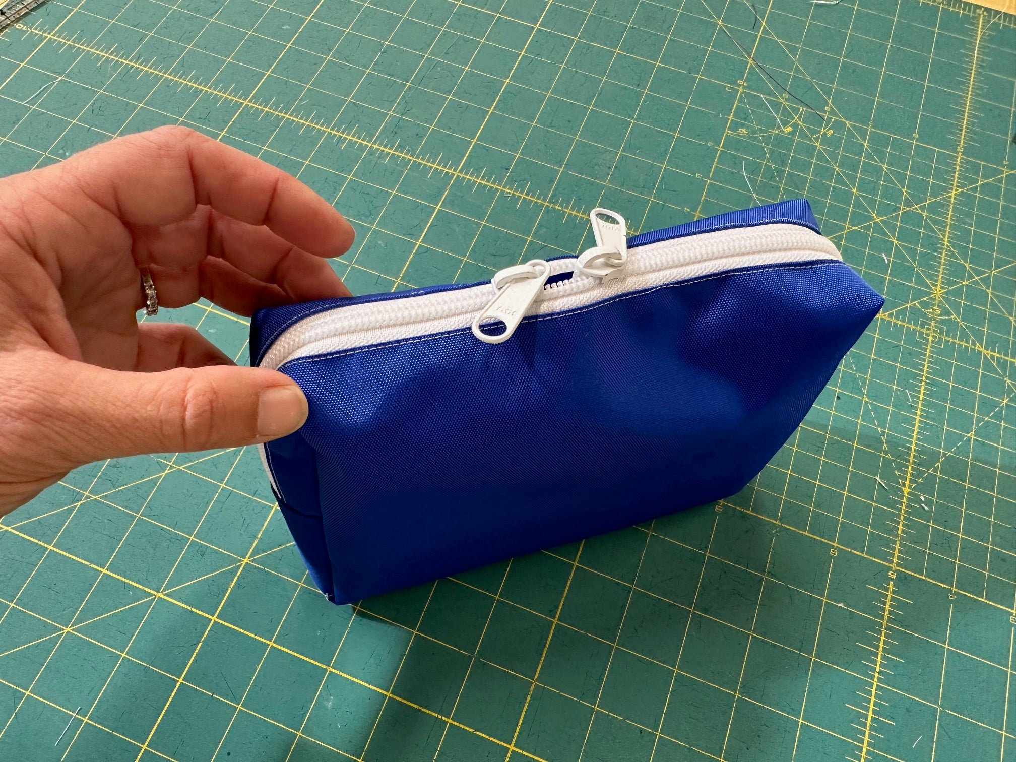 Blue zippered pouch with white zipper held by a hand on a green cutting mat.
