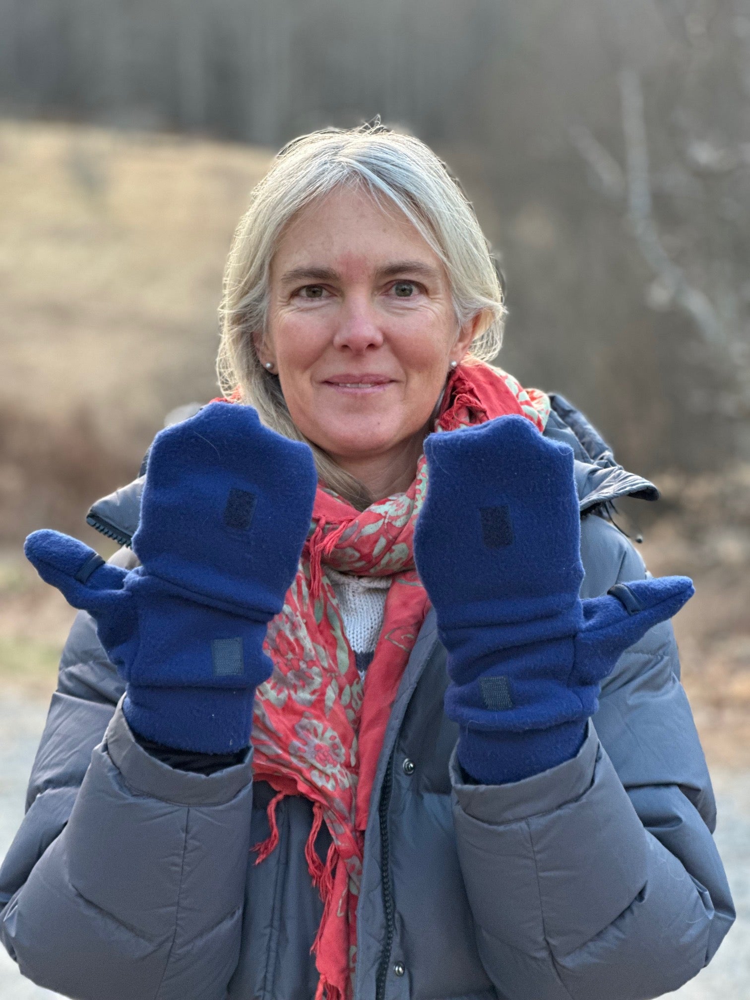 Woman wearing blue mittens and a gray coat outdoors with a blurred natural background