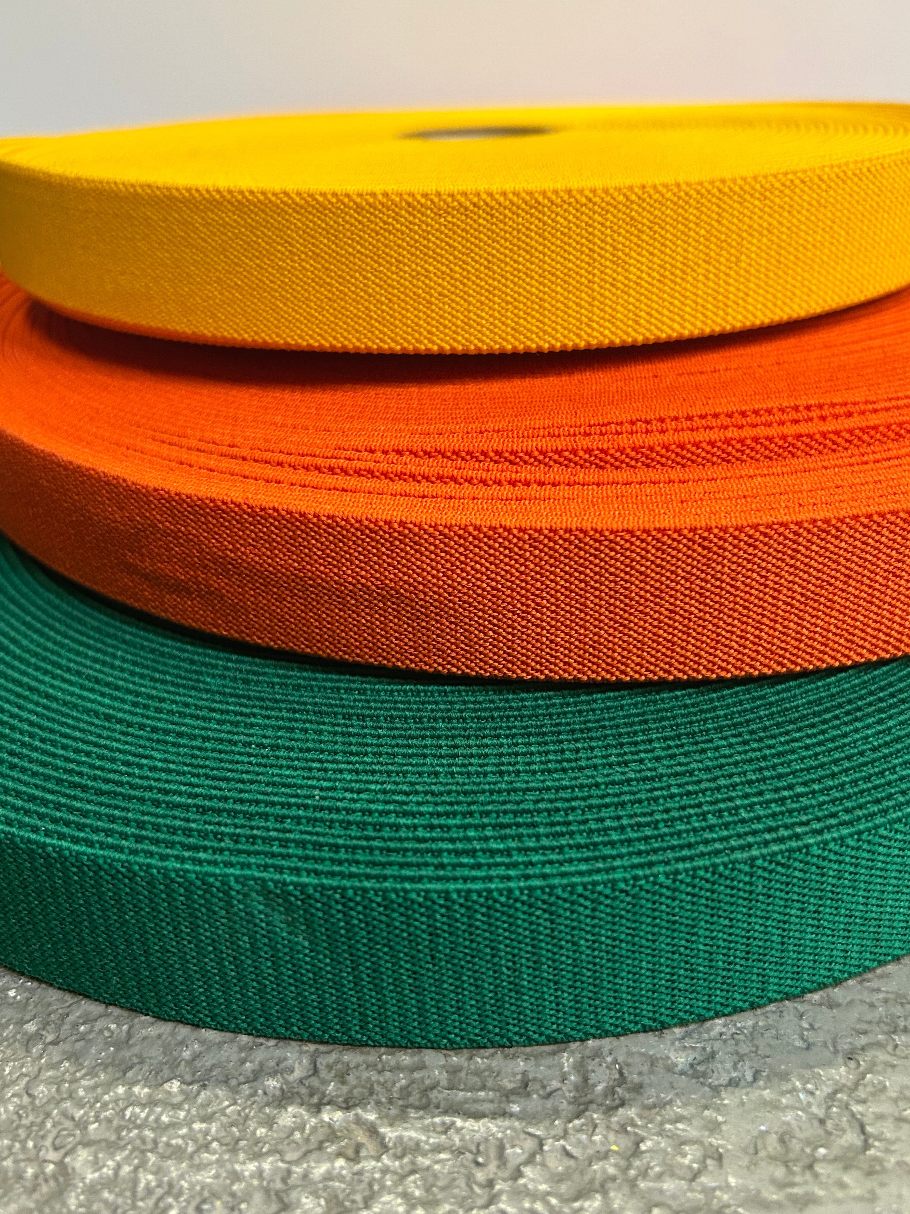 Three rolls of webbing elastic in yellow, orange, and green on a textured surface.