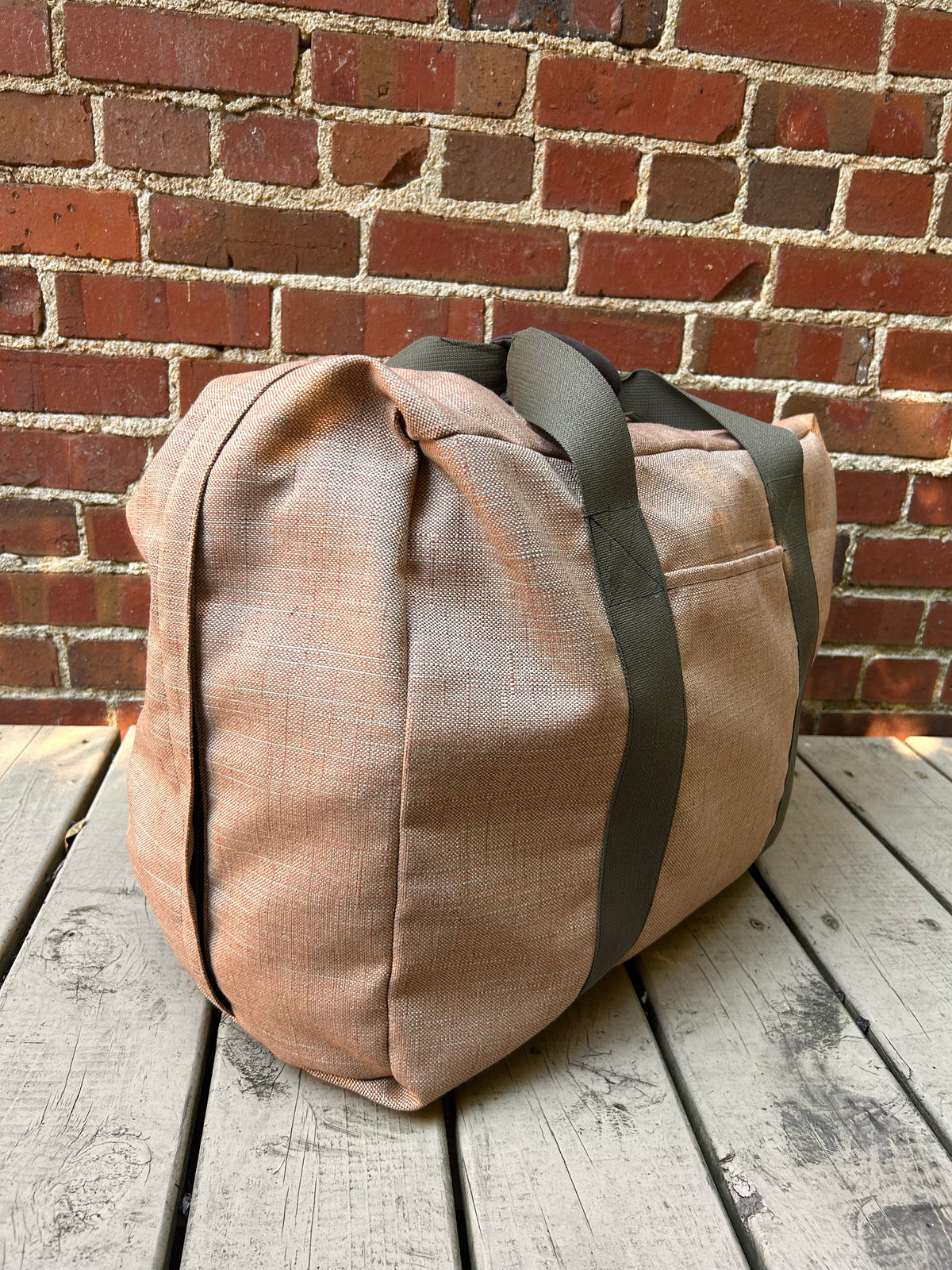 soft fabric suitcase in brown with olive webbing handles on a wooden deck and against a brick wall.