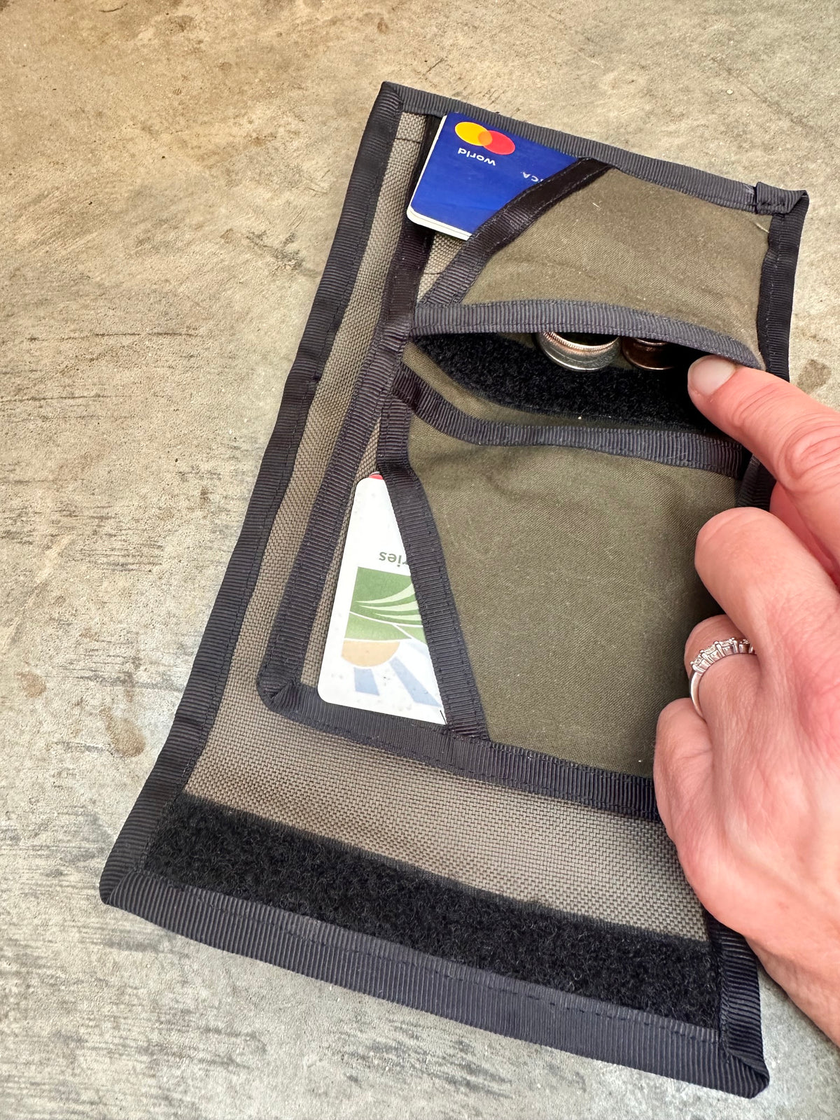 Small olive colored wallet with black trim opened up on a concrete floor -- with cards and change.