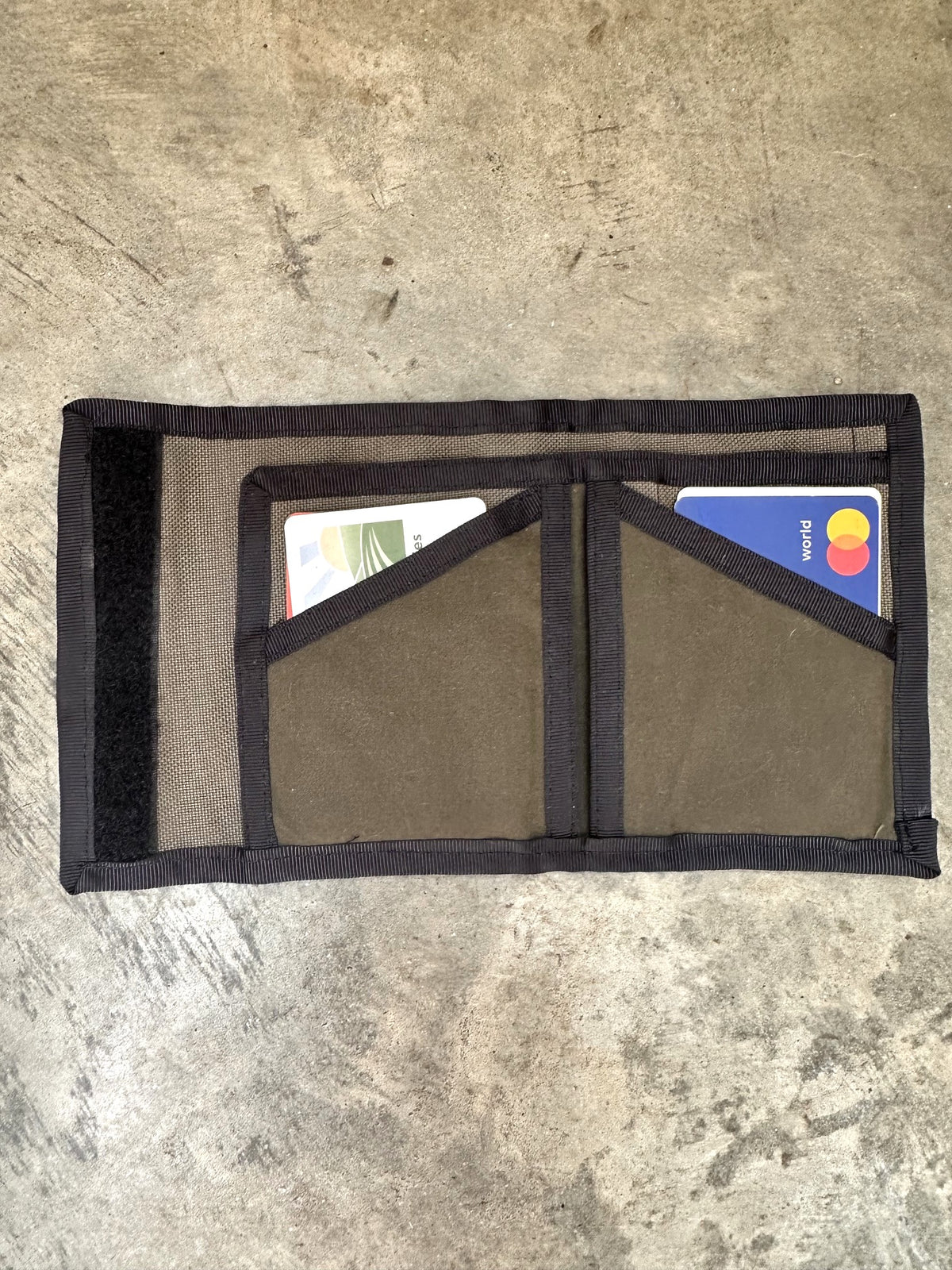 Small olive colored wallet with black trim opened up on a concrete floor