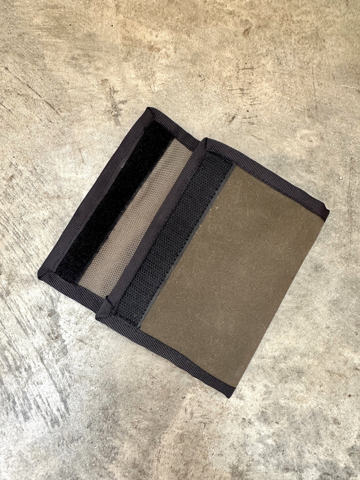 Small olive colored wallet with black trim opening up on a concrete floor