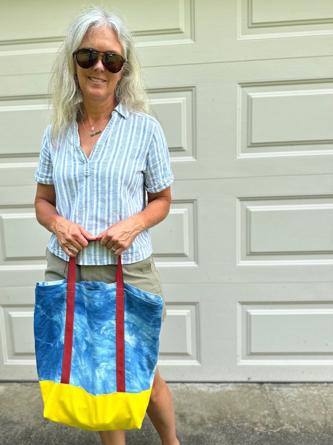 A woman with gray hair and sunglasses stands in front of a white garage door holding a blue and yellow tie-dye tote bag with red handles.