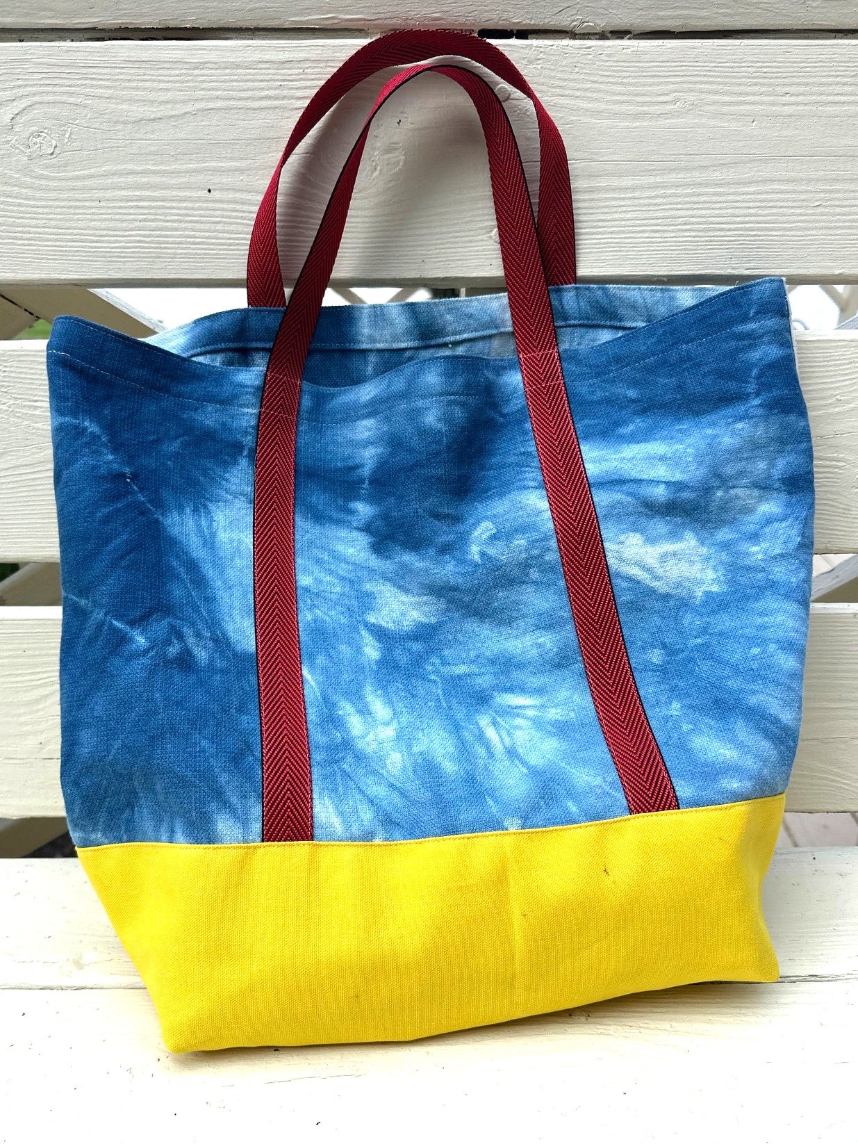 A handmade tote bag with a blue and white tie-dye pattern on the upper half, a solid yellow bottom panel, and dark red webbing handles, resting on a white wooden slatted surface.