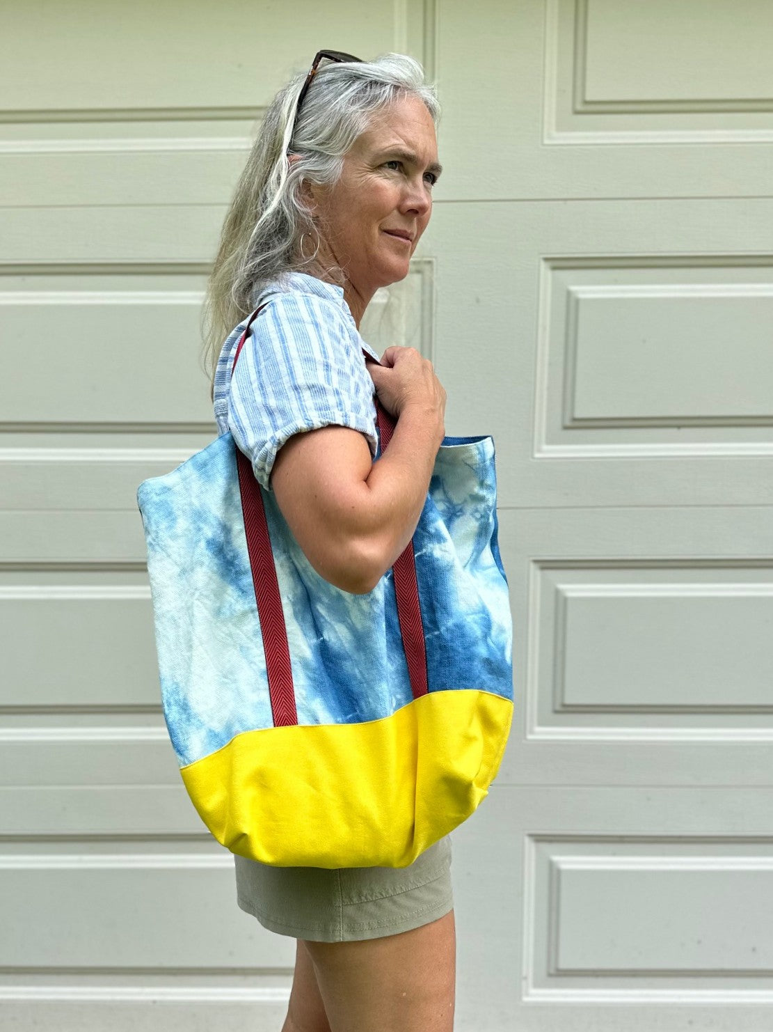 A woman with gray hair wearing a blue and white striped shirt and khaki shorts carries a large tie-dye tote bag with blue and yellow panels and red straps, standing in front of a white garage door.