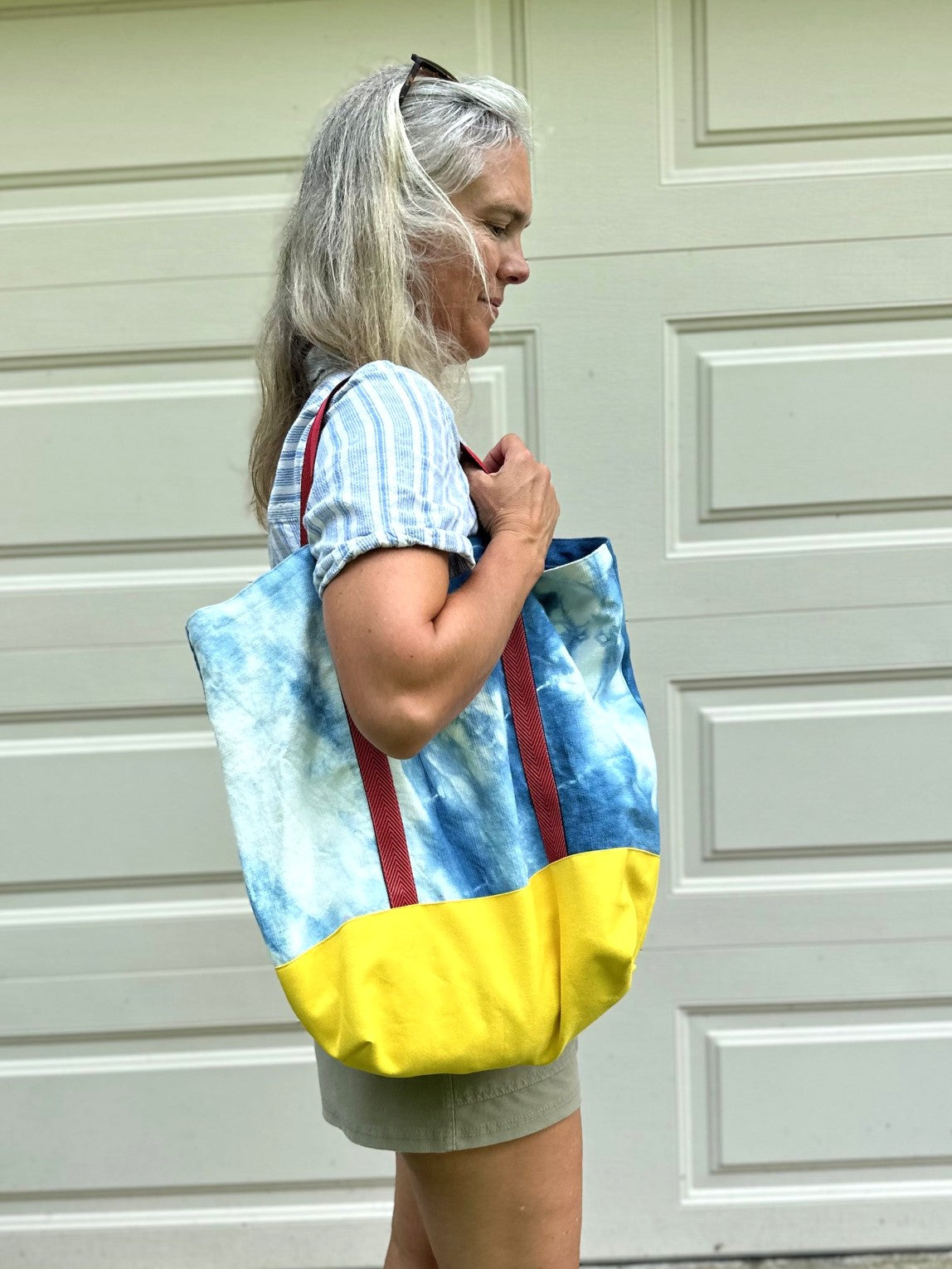 A woman with gray hair tied back is shown from the side, wearing a blue and white tie-dye tote bag with a solid yellow bottom and red straps, standing in front of a white garage door.