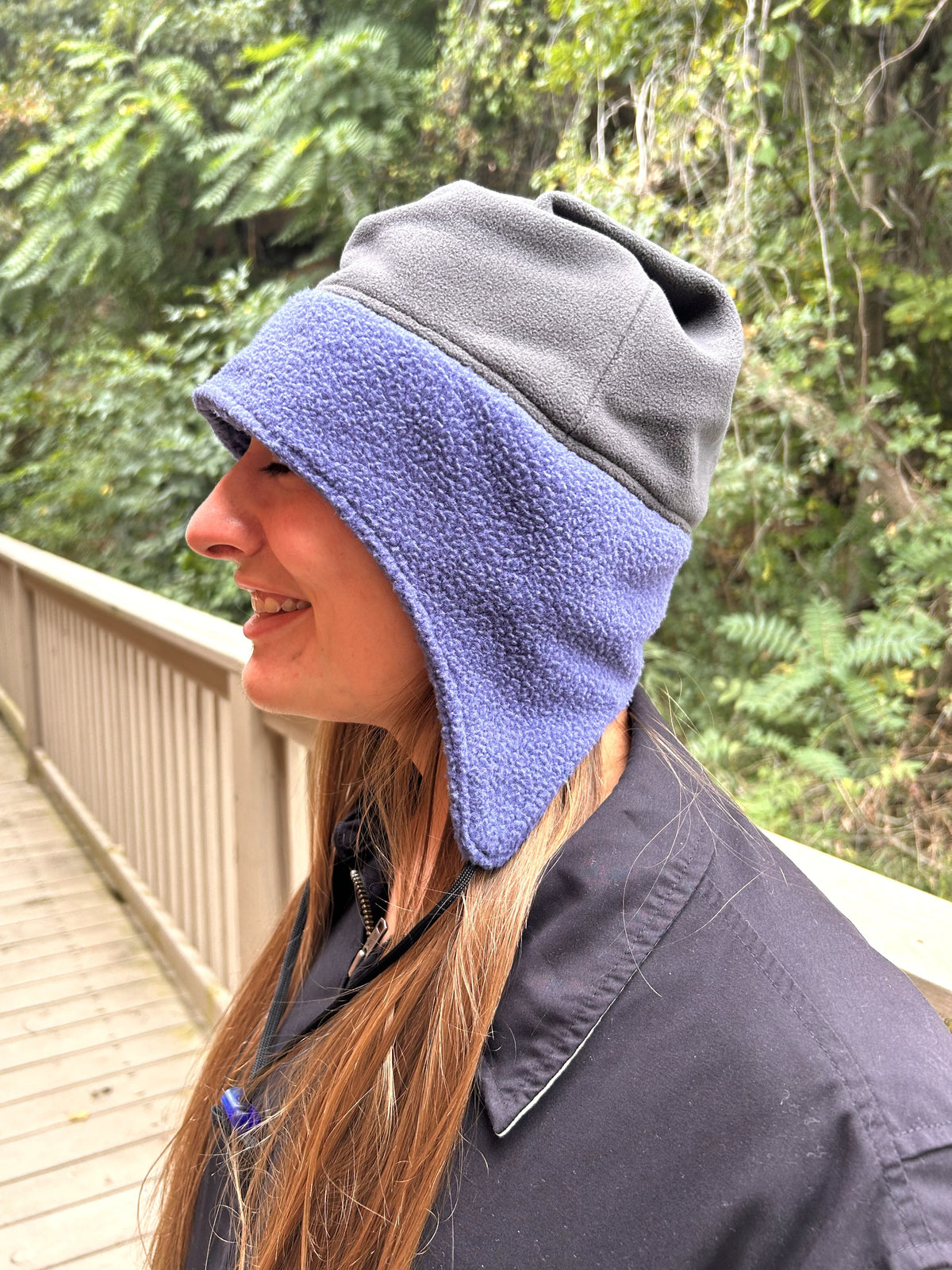 Woman standing outside by a fence and trees wearing a dark jacket and a two-color (blue and grey) toboggan. Side facing camera