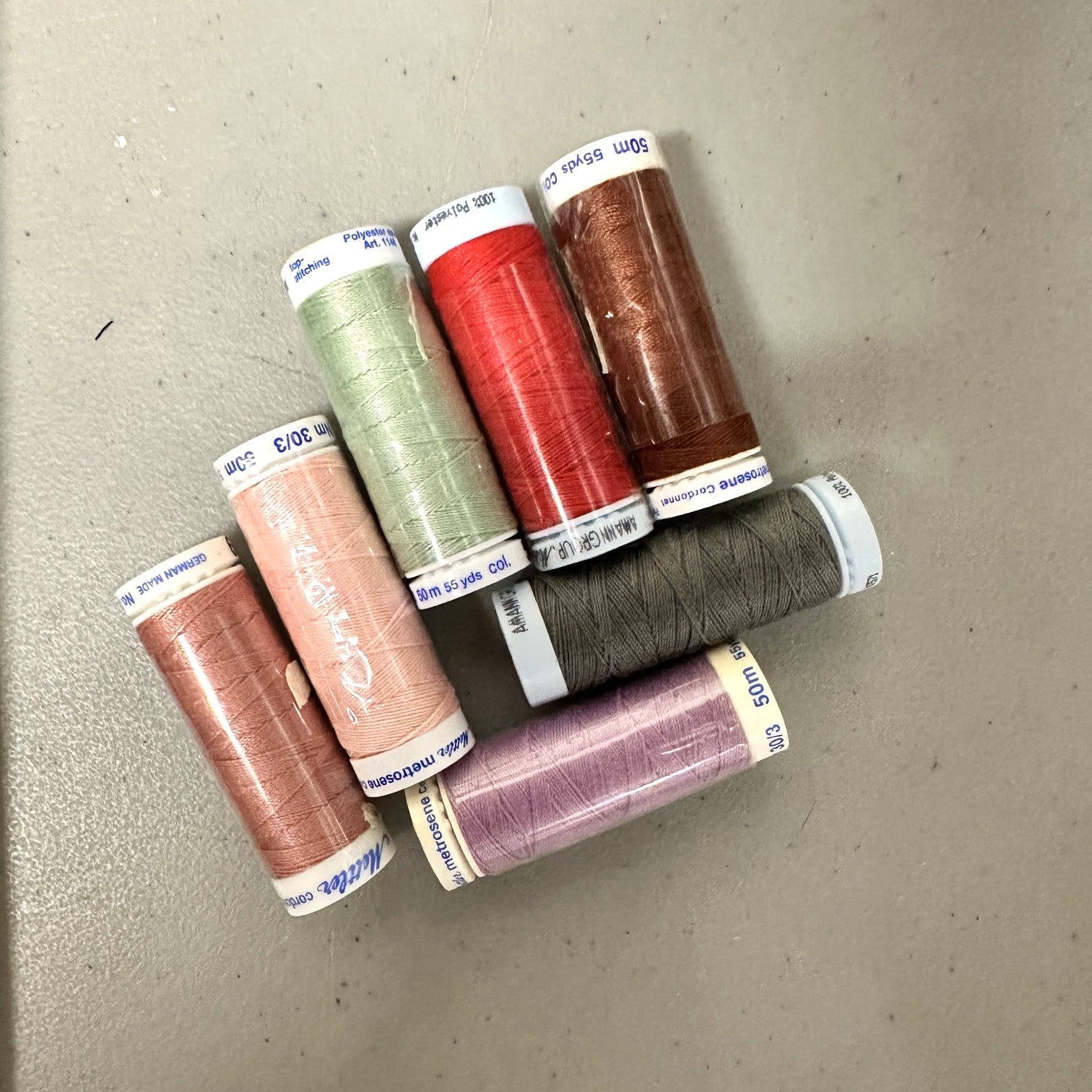 Seven spools of sewing thread in assorted colors, including pink, green, red, brown, gray, and purple, arranged on a neutral gray surface.
